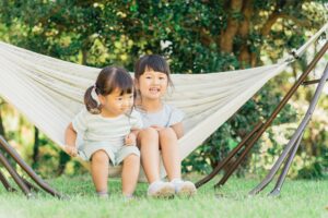 Two young girls sitting in a white hammock outdoors with green trees in the background.