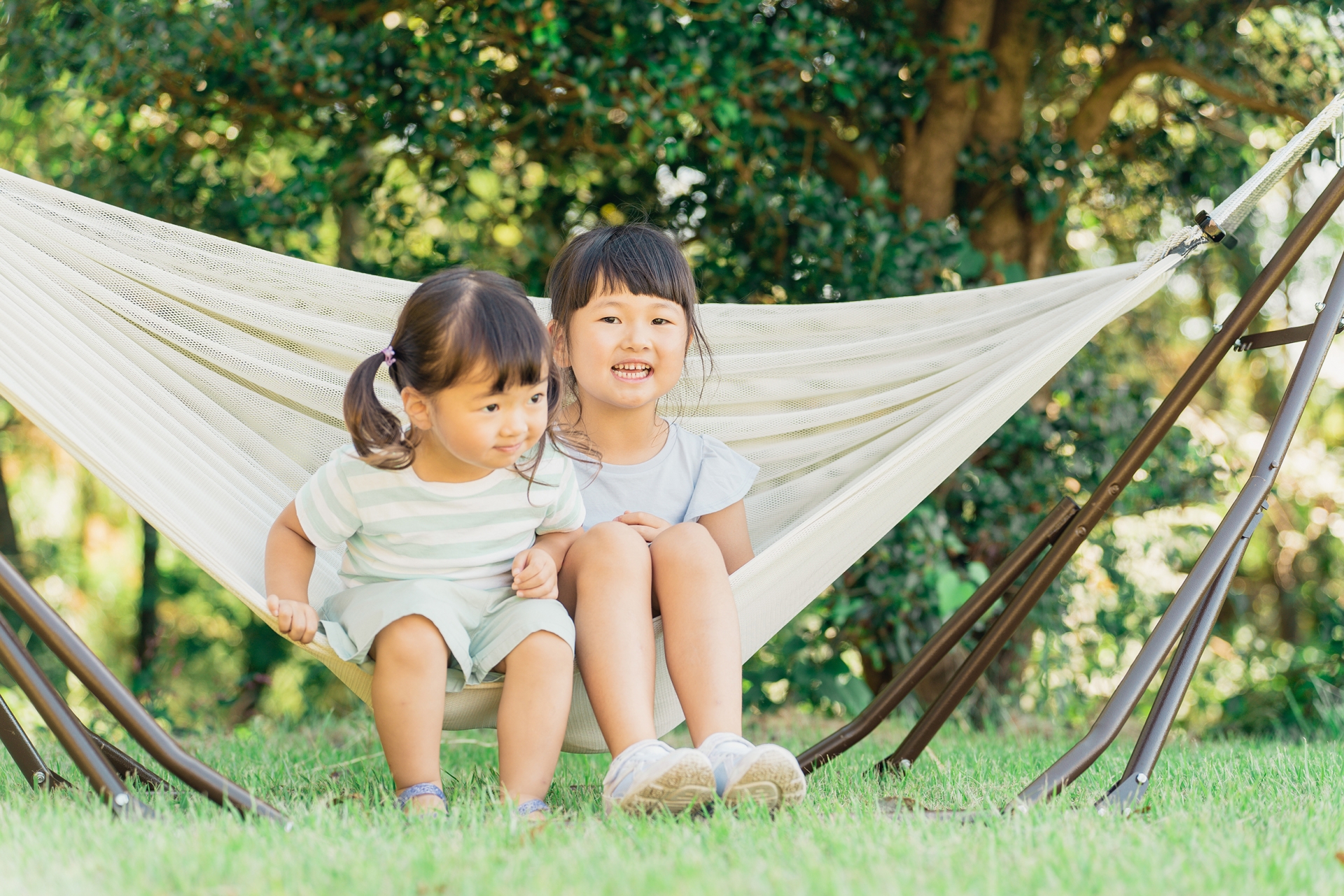 Two young girls sitting in a white hammock outdoors with green trees in the background.