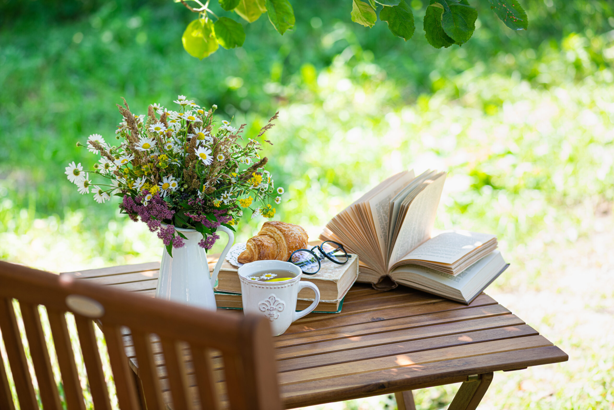 A bouquet of wildflowers in a white pitcher on a wooden table with croissants, books, a cup of tea, and eyeglasses outdoors in a sunny garden.