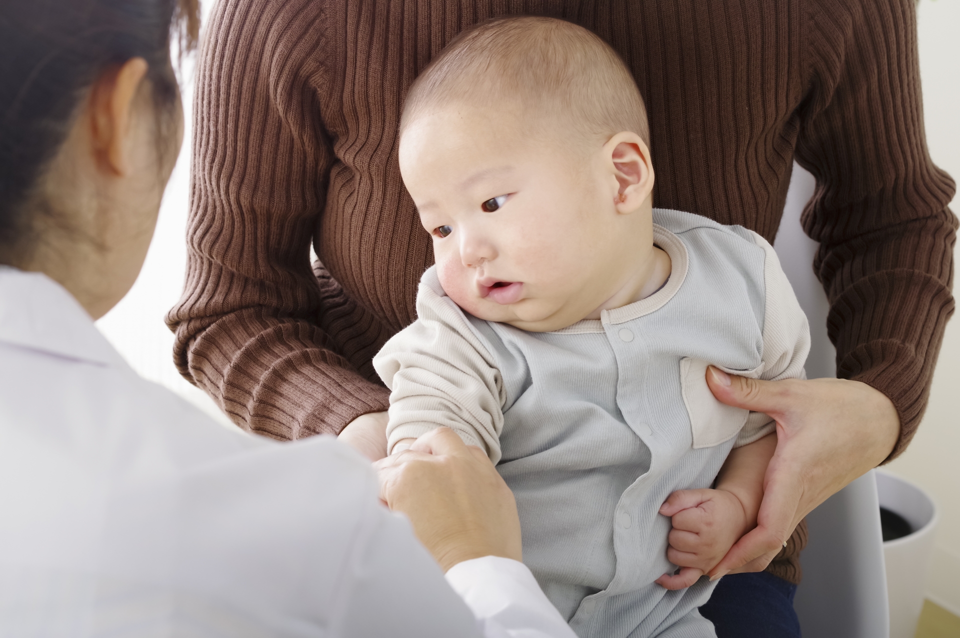 Infant in a light gray onesie held by a caregiver during a medical checkup with a clinician nearby