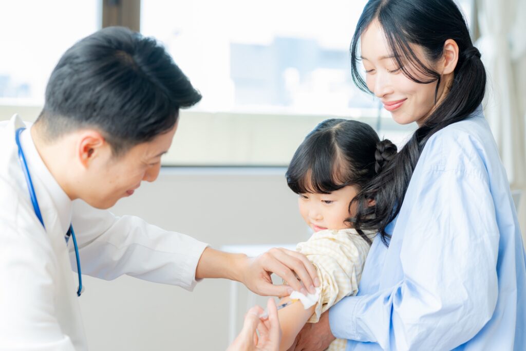 Mother holding a young girl while a healthcare professional administers a vaccine injection to the child's arm, in a clinical setting.