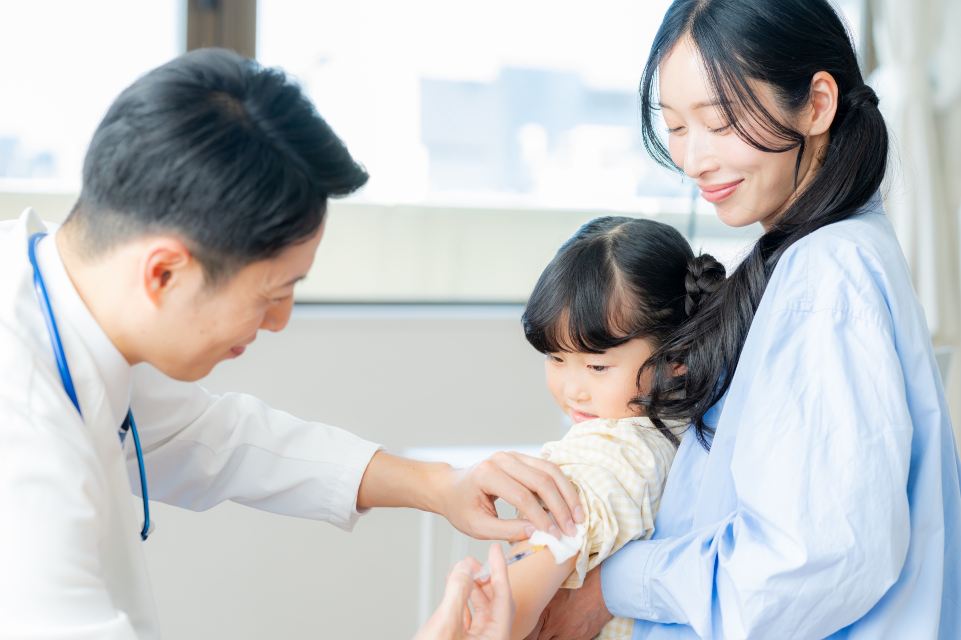 Mother holding a young girl while a healthcare professional administers a vaccine injection to the child's arm, in a clinical setting.