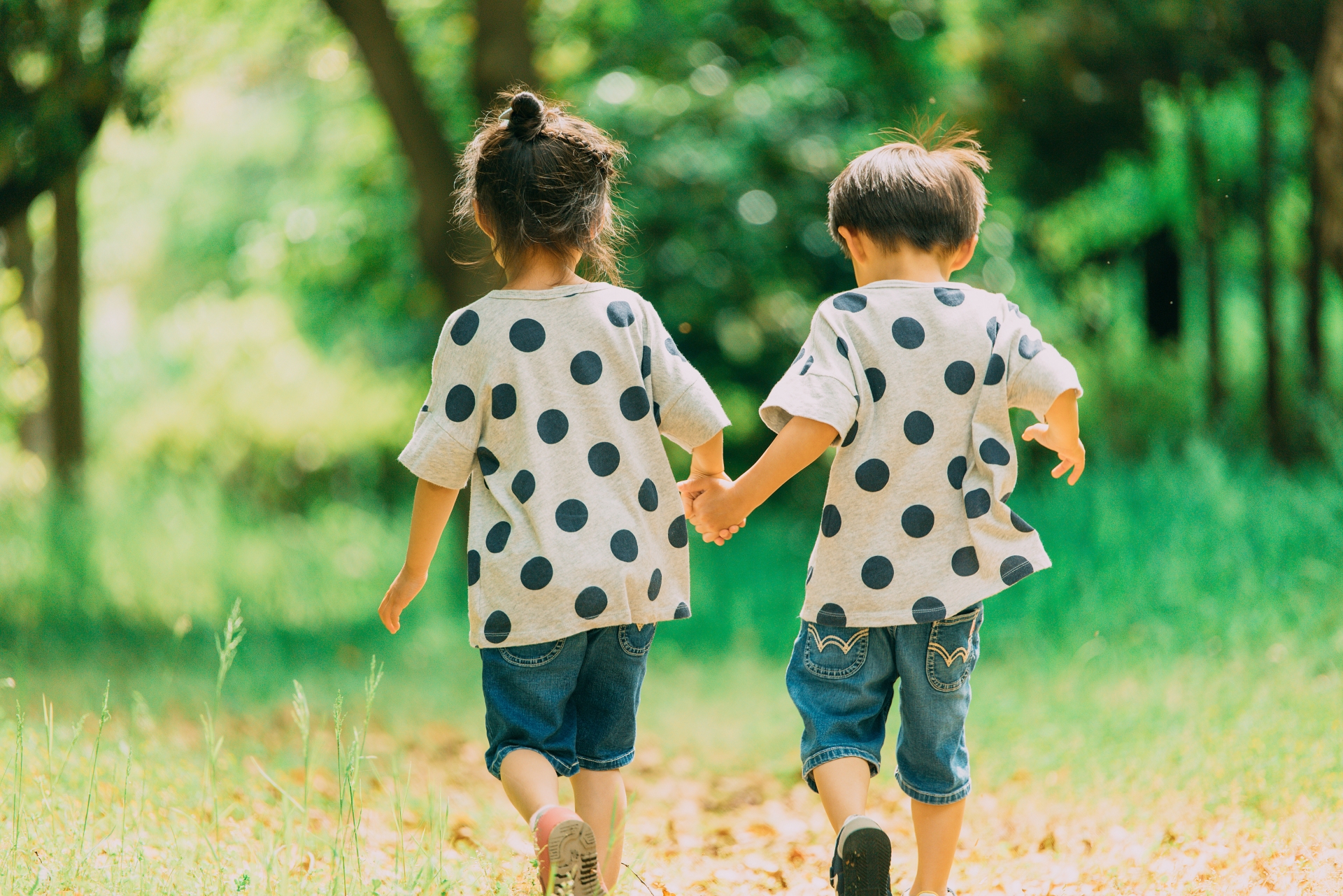 Two young children hold hands and walk away from the camera along a sunlit forest path, wearing gray polka-dot shirts and jeans.